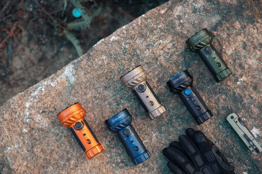 Mehrere Olight Taschenlampen in Orange, Blau, Grün auf einem Felsen, daneben Outdoor-Handschuhe und Messer.
