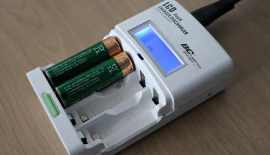 Batterieladegerät mit zwei eingelegten AA-Batterien auf einem Holztisch. LCD-Display zeigt Ladevorgang an.