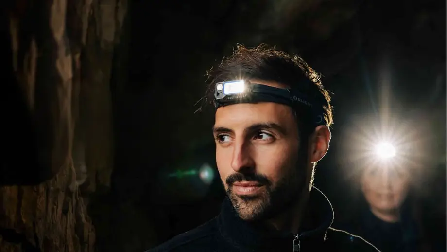 Mann mit leuchtender Stirnlampe in dunkler Höhle, zweiter Lichtkegel im Hintergrund.