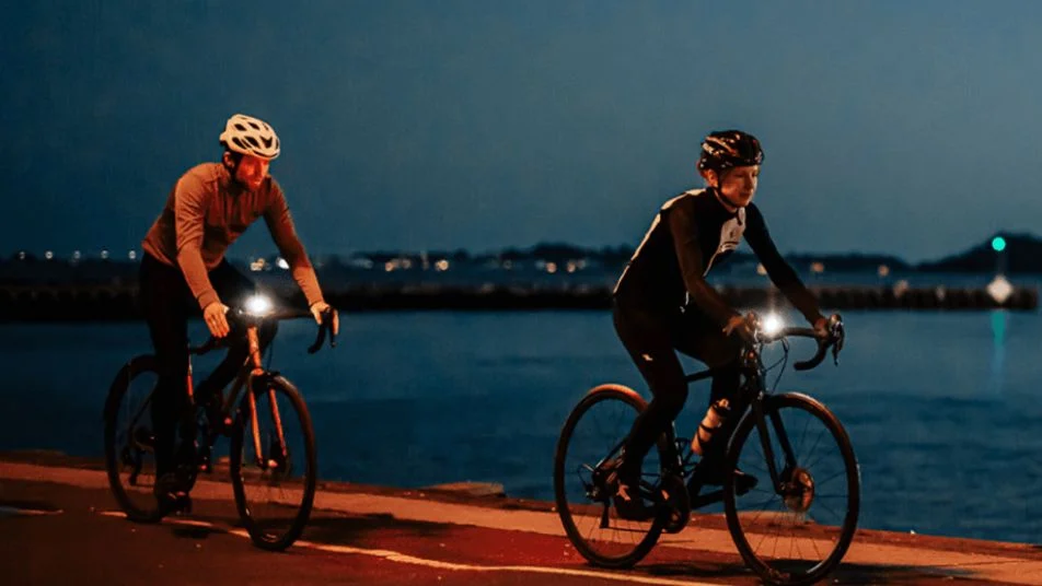 Zwei Radfahrer nachts am Wasser mit hellen Fahrradlichtern unterwegs.