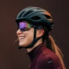Lächelnde Rennradfahrerin mit schwarzem Kask Helm und verspiegelter Sportbrille im Profil vor dunklem Hintergrund.