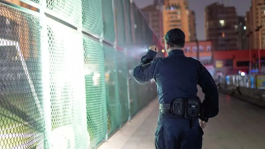 Wachmann leuchtet nachts mit Taschenlampe auf einen grünen Zaun in der Stadt.