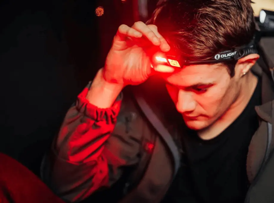 Mann mit Stirnlampe, die rotes Licht ausstrahlt, in dunkler Umgebung. Er trägt eine Jacke und konzentriert sich auf das Licht.
